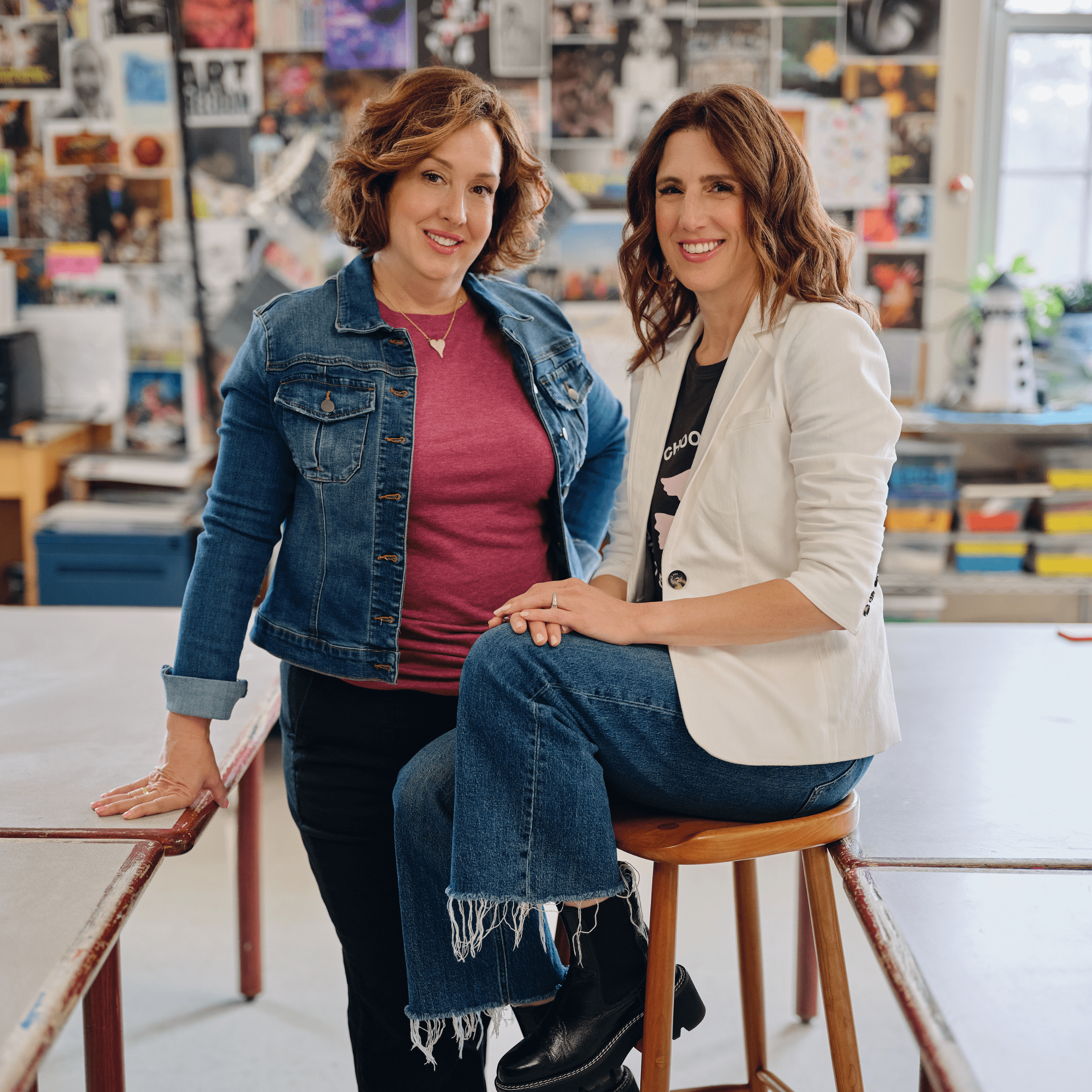 Drs. Kristie and Julie pose in a school classroom, standing in front of a vibrant wall filled with art, magazine clippings, and colorful storage bins. The lively background underscores their commitment to inclusive schooling, promoting creativity and equitable learning environments for all students.