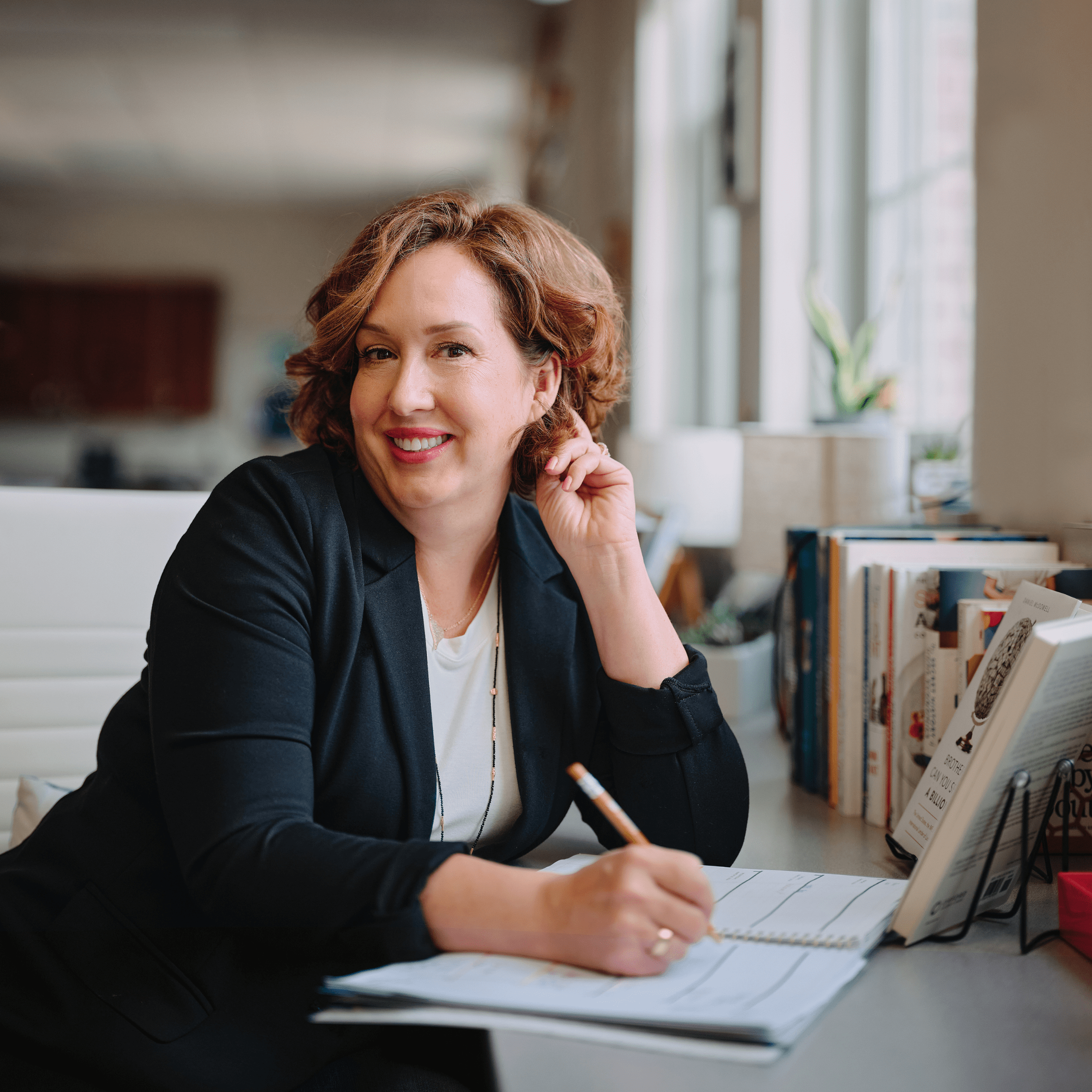 Dr. Julie Causton writes in her planner at a desk, wearing a black blazer. Her focused expression highlights her commitment to organizing and implementing inclusive strategies in education.