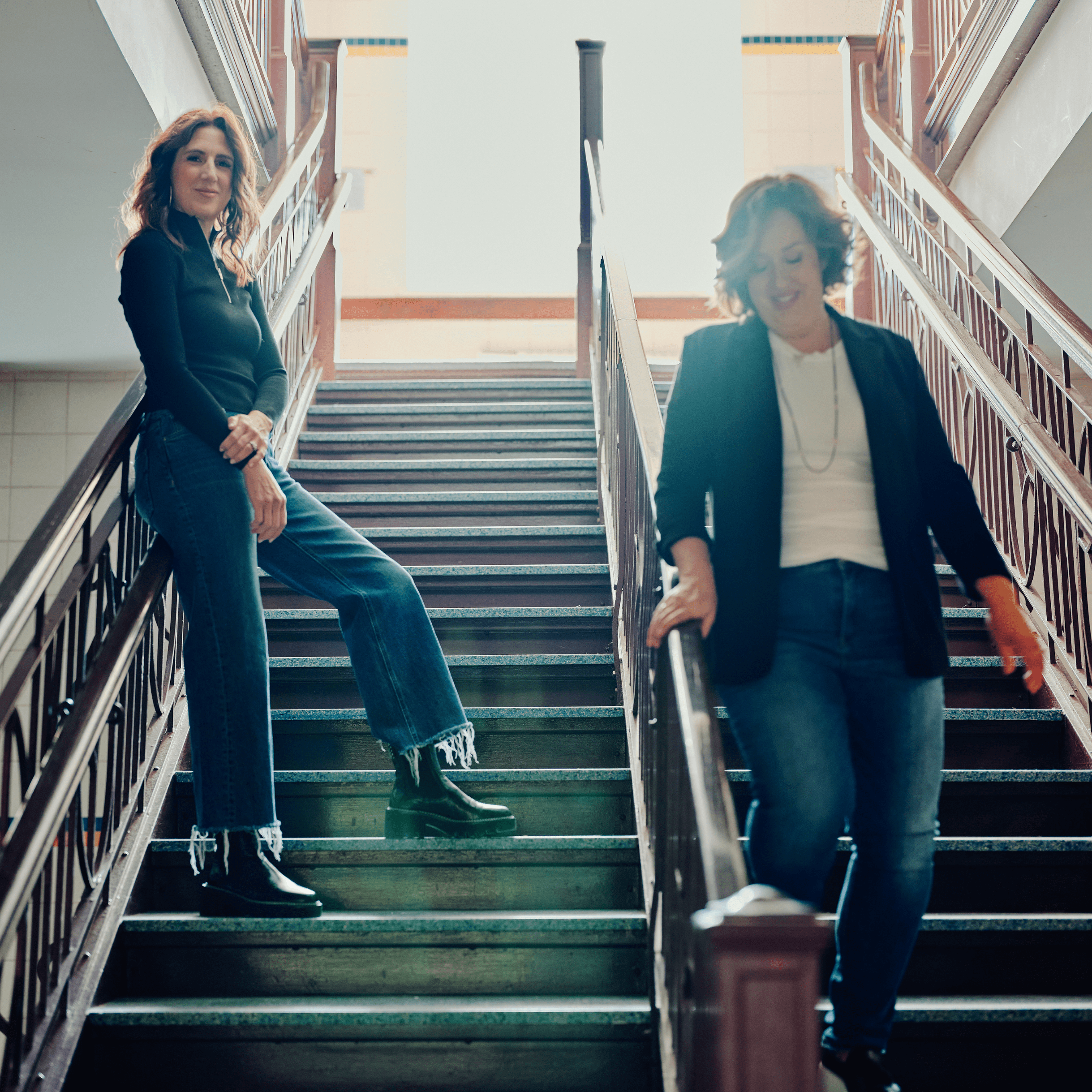 Drs. Kristie and Julie walk down a school stairway with light streaming in from a window behind them. Both are smiling and laughing, dressed in professional attire, capturing a moment of joy and camaraderie.