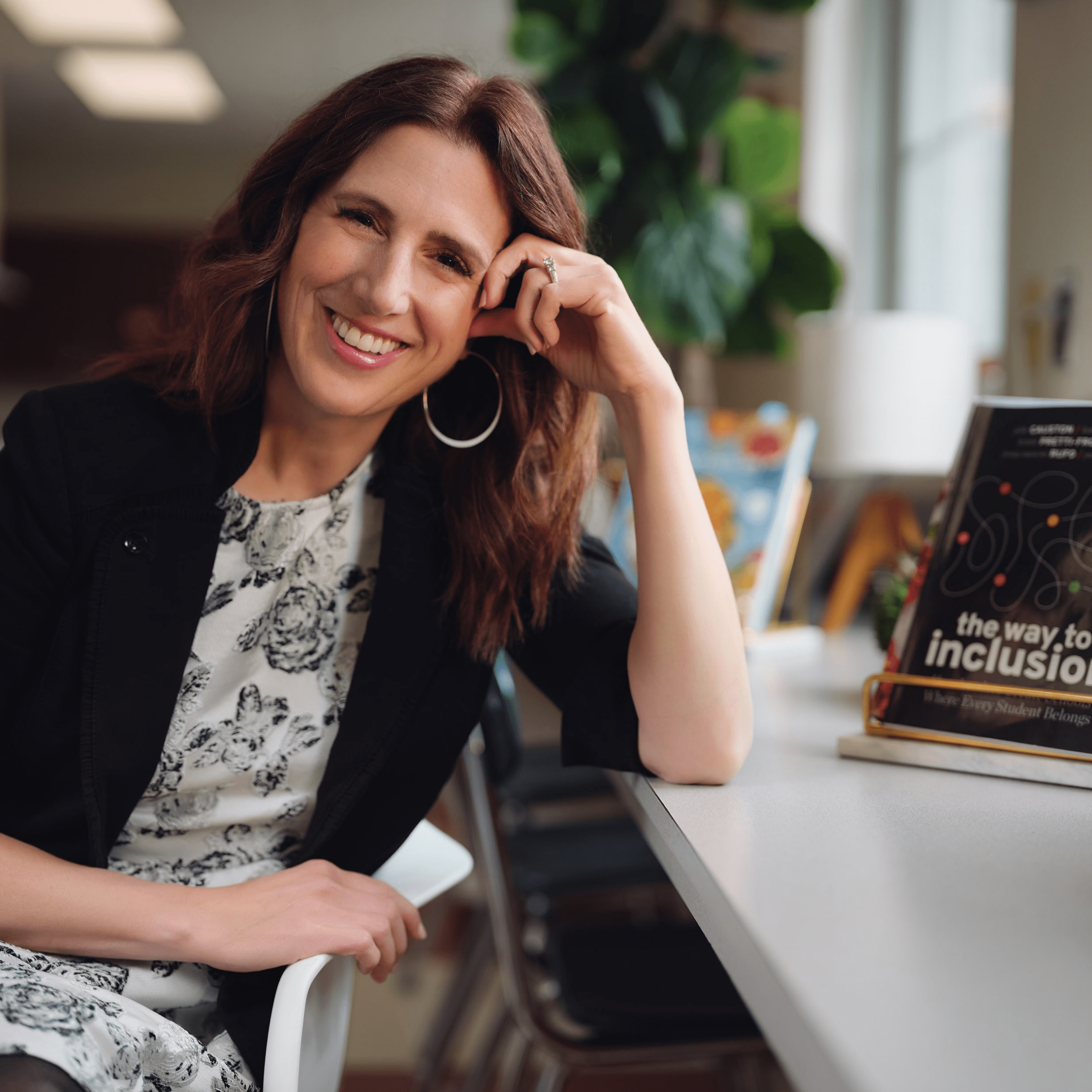 Dr. Kristie smiles at the camera while seated and leaning on a desk behind her. The book 'The Way to Inclusion' is visible on the desk, with a window in the background, creating a warm and welcoming atmosphere.