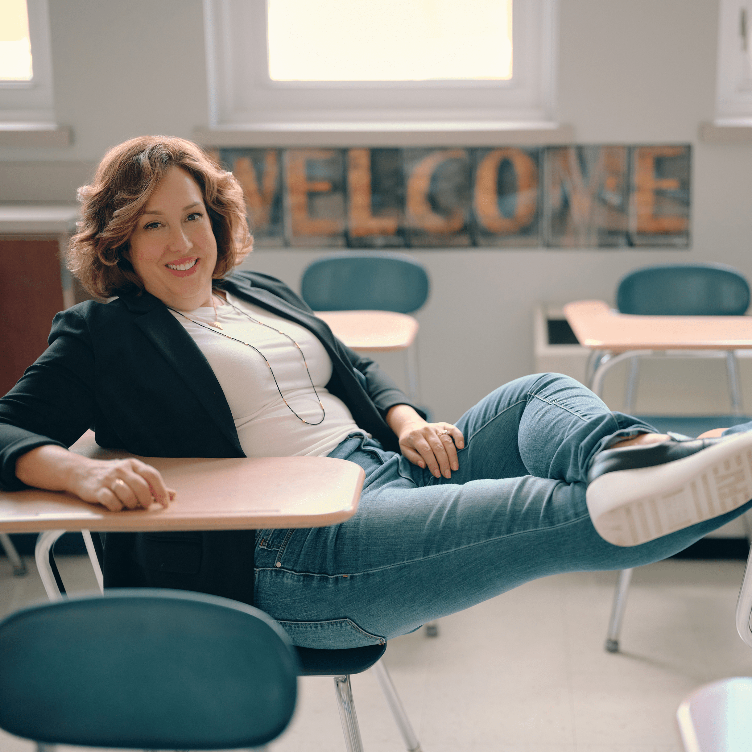 Dr. Julie lounges comfortably in a school desk, wearing a professional blazer, jeans, and fun sneakers. A welcome sign behind her emphasizes her commitment to fostering an inclusive and inviting atmosphere in educational settings.